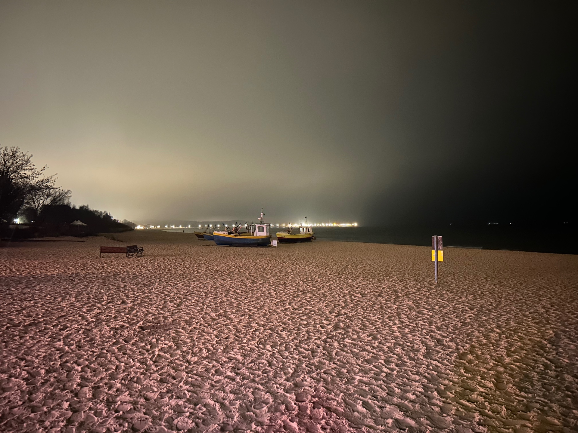 Łodzie rybackie na plaży nocą - klimatyczny blask odległych świateł miasta odbijający się na spokojnym morzu